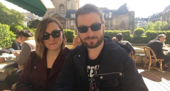 Aura und Bogdan sitzen in einem Café oder Restaurant, im Hintergrund ist eine Kirche zu sehen. Die Sonne scheint, die beiden tragen Sonnenbrillen und lächeln selbstironisch-souverän in die Kamera.