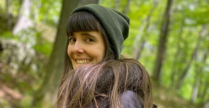 Julia steht im Wald und schaut lächelnd in die Kamera.