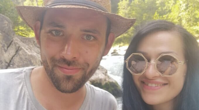 Vicky und Sebastian stehen nebeneinander und lächeln in die Kamera. Sebastian hat einen Strohhut auf und Vicky trägt eine coole 8-eckige Sonnenbrille. Im Hintergrund sind Wald und ein Fluß zu sehen.